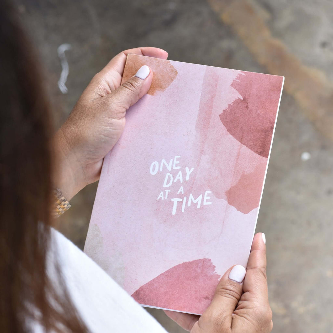 One Day at a Time Journal with pink watercolor background and white text held in hands, suitable as a sympathy gift.
