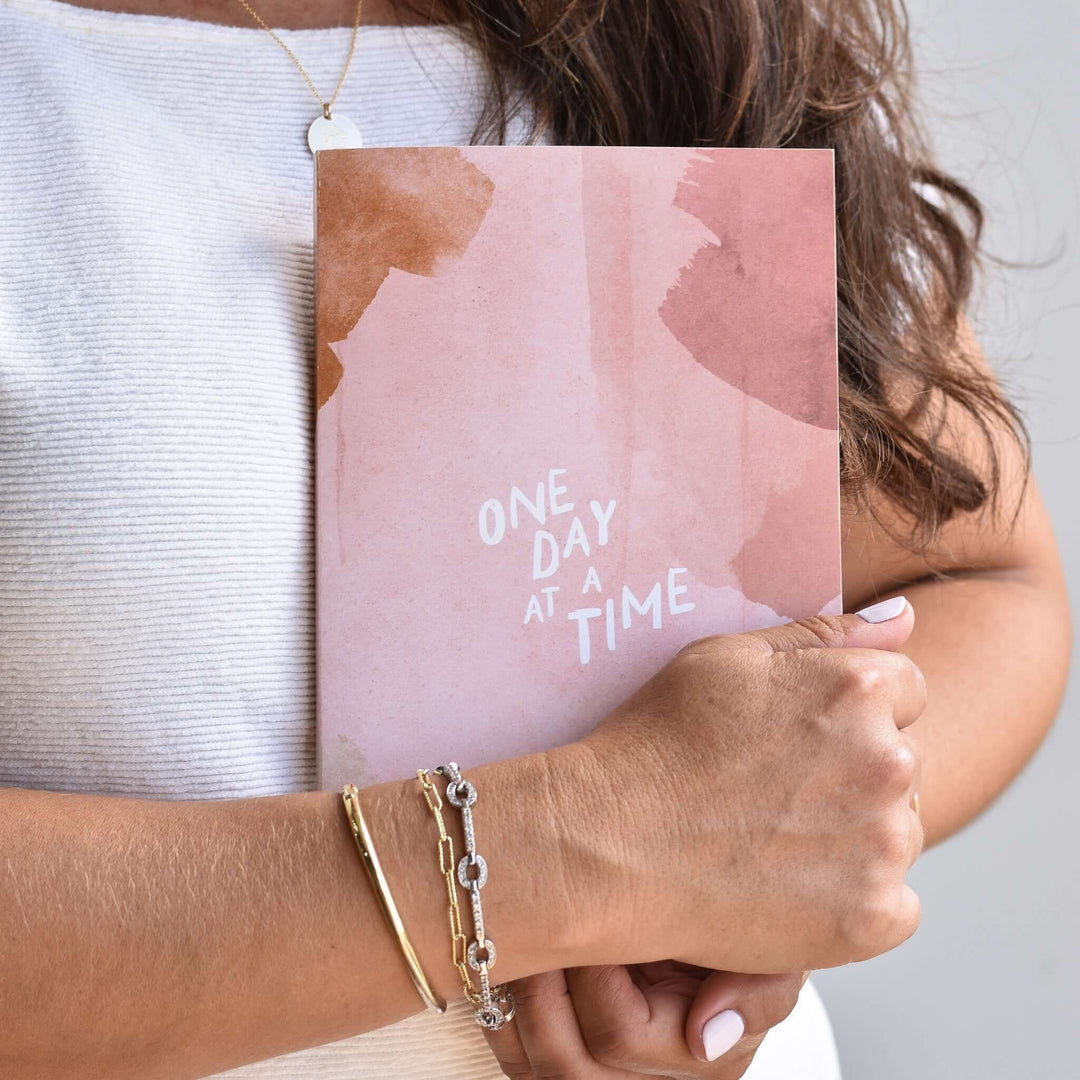 One Day at a Time Journal held by a person, featuring a pink cover with abstract art and white text.