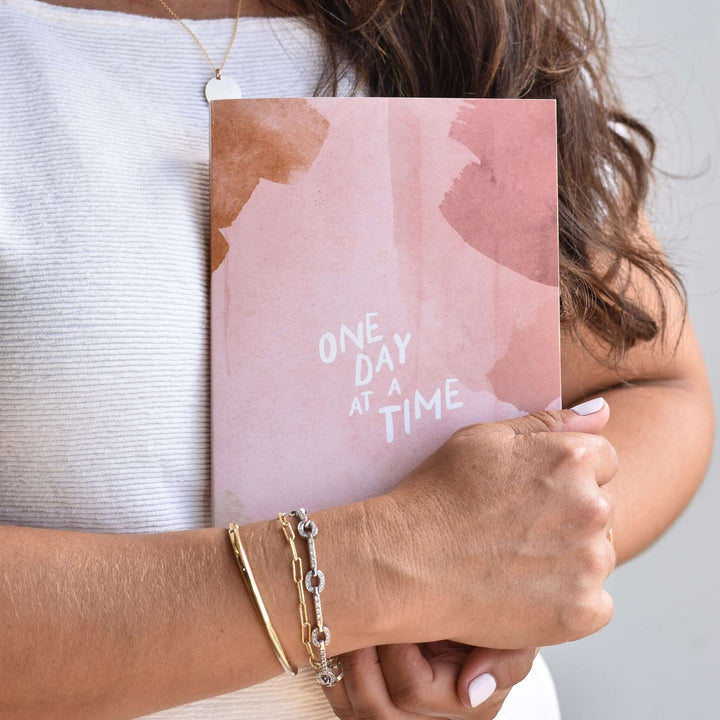 One Day at a Time Journal held by a person, featuring a pink cover with abstract art and white text.