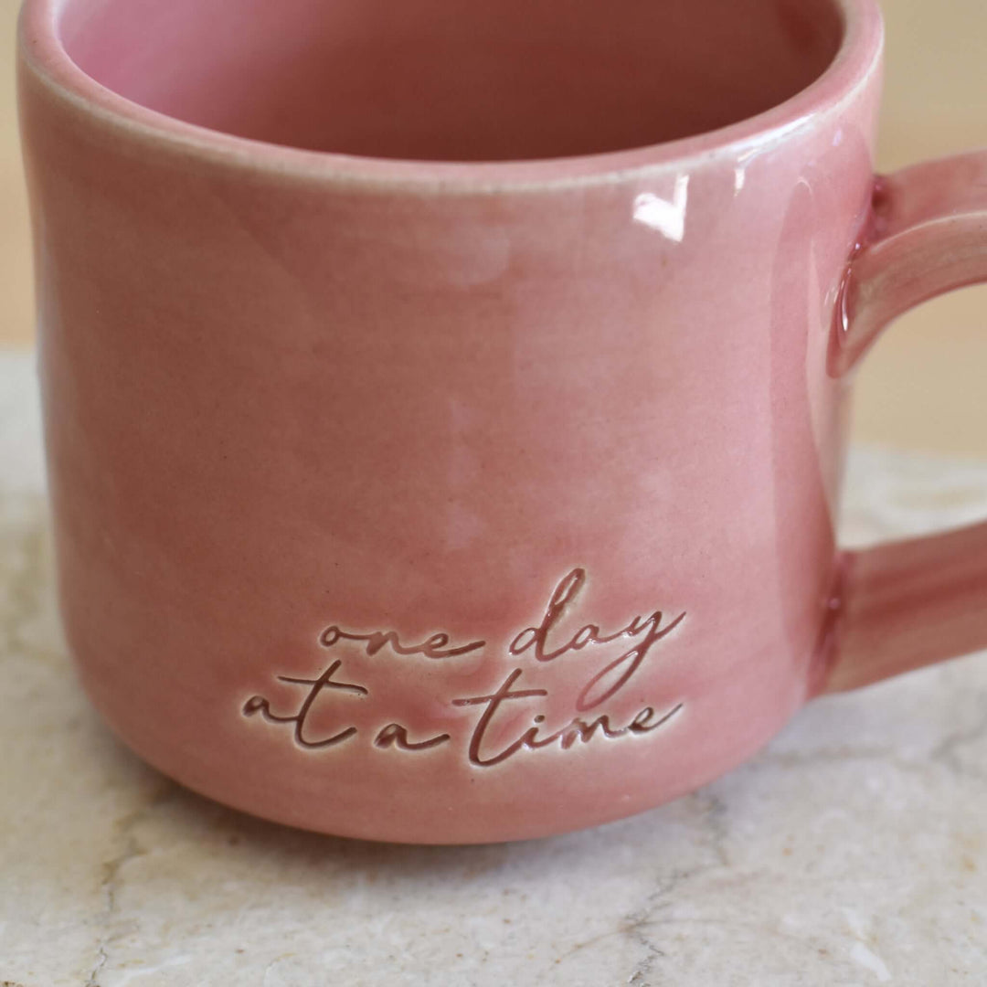One Day at a Time Mug in pink ceramic with embossed text, shaped with a handle, resting on a light-colored surface.