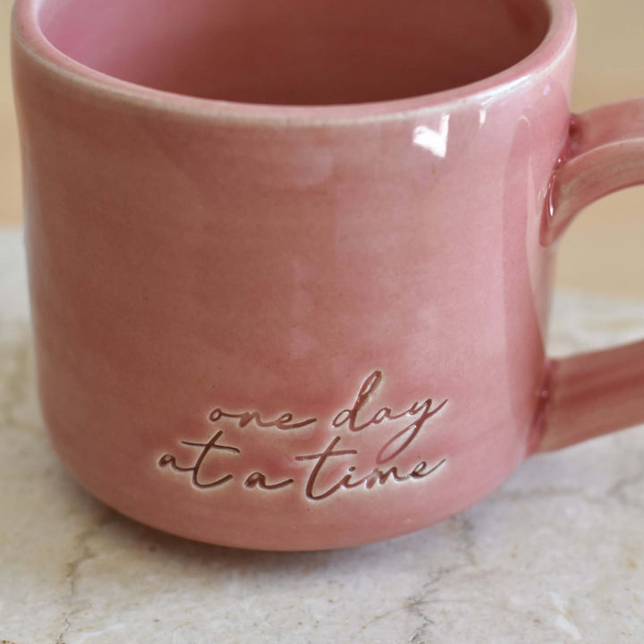 One Day at a Time Mug in pink ceramic with embossed text, shaped with a handle, resting on a light-colored surface.