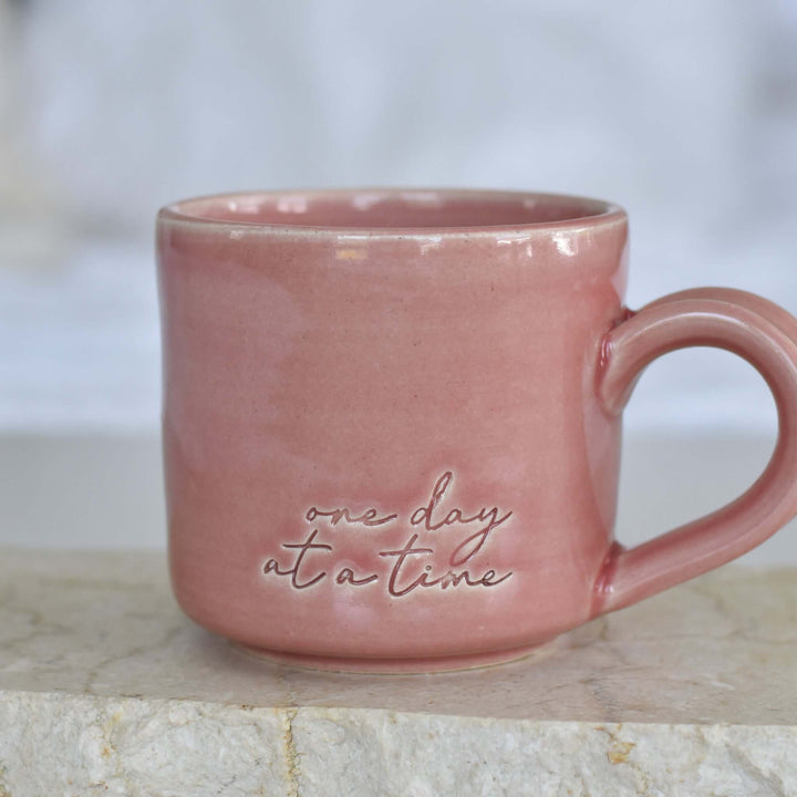 One Day at a Time Mug, pink ceramic, round shape, engraved text, smooth surface, placed on a light stone background.