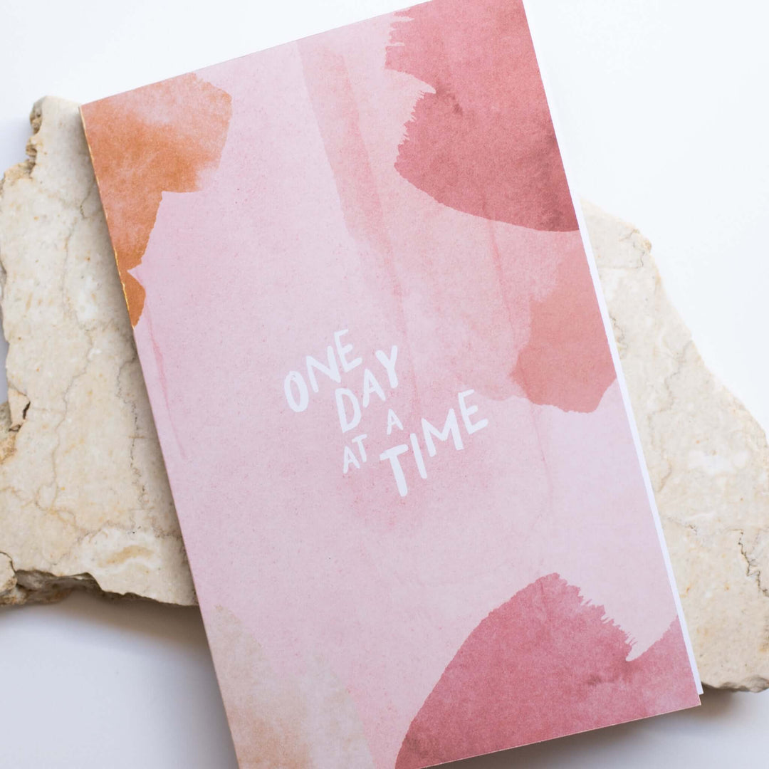 One Day at a Time Gift Box notebook with pink watercolor design and white text on top, placed on a stone surface.