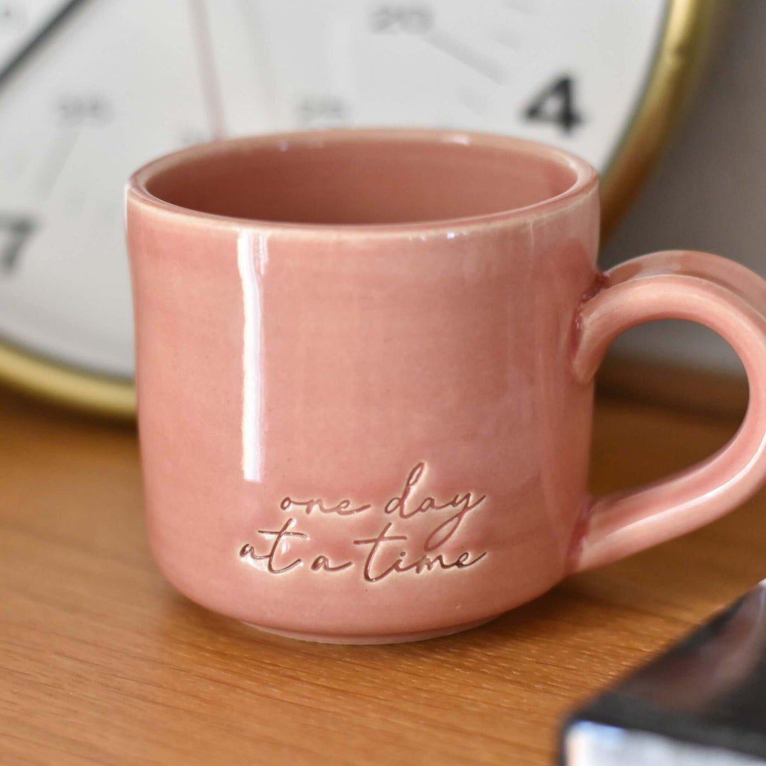 One Day at a Time Mug in pink ceramic with embossed text, placed on a wooden surface near a clock.