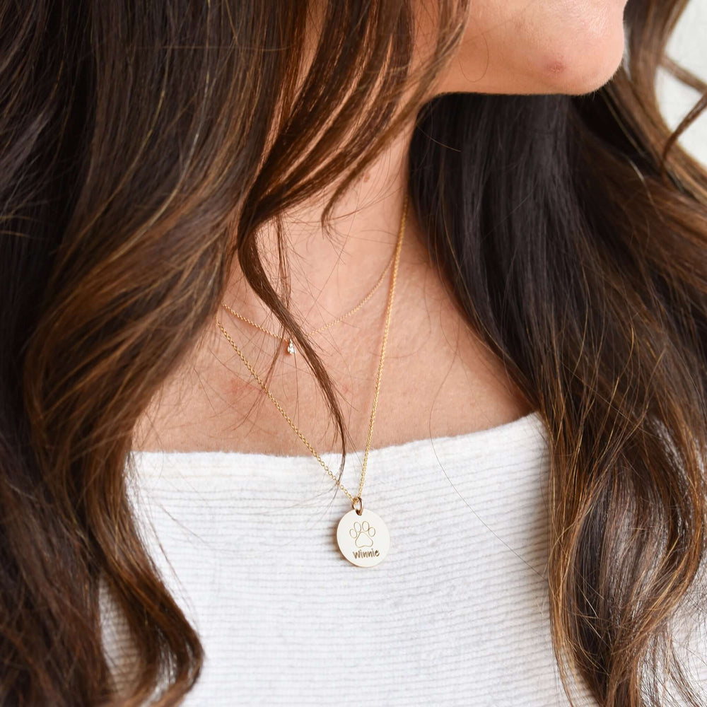 Personalized Pet Necklace in gold with engraved name 'Winnie' and paw print design, worn by a person with dark hair.