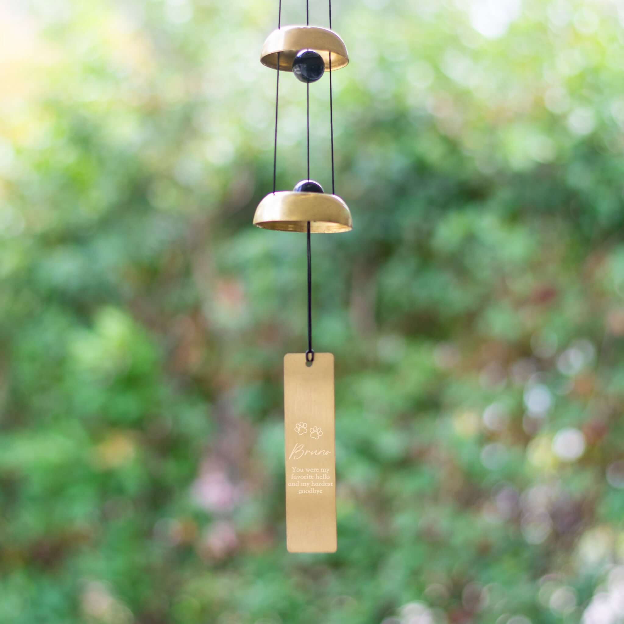 Pet Memorial Wind Chime with two gold bells and a rectangular engraved tag, hanging against a blurred green background.