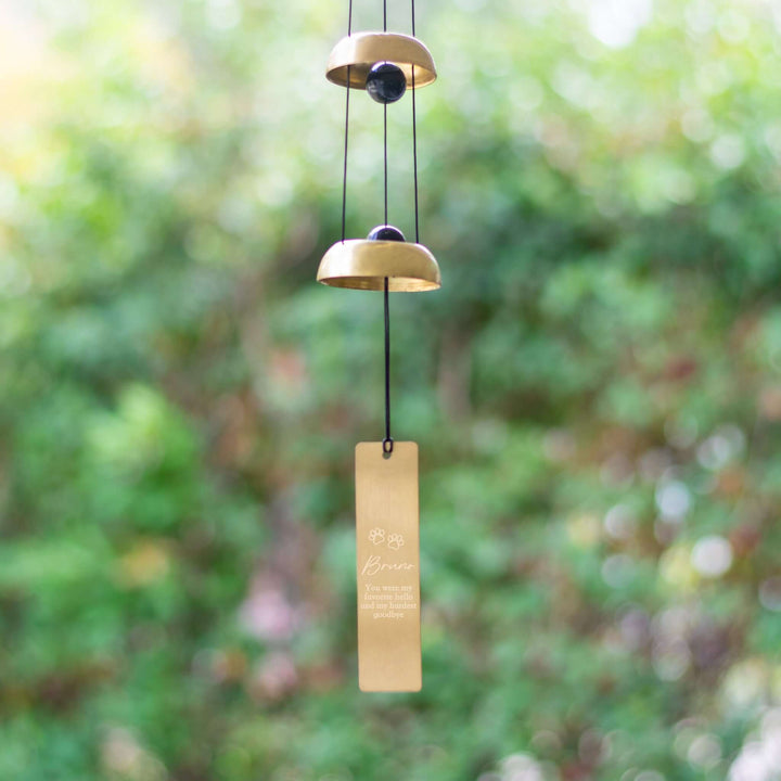 Pet Memorial Wind Chime with two gold bells and a rectangular engraved tag, hanging against a blurred green background.