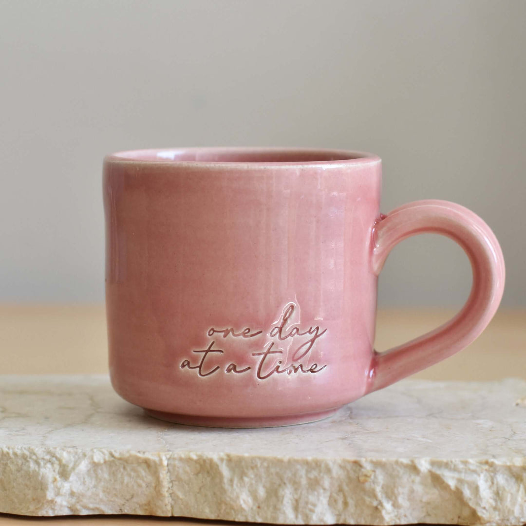 One Day at a Time Mug, pink ceramic mug with hand-glazed finish, features embossed text, sitting on a stone surface.