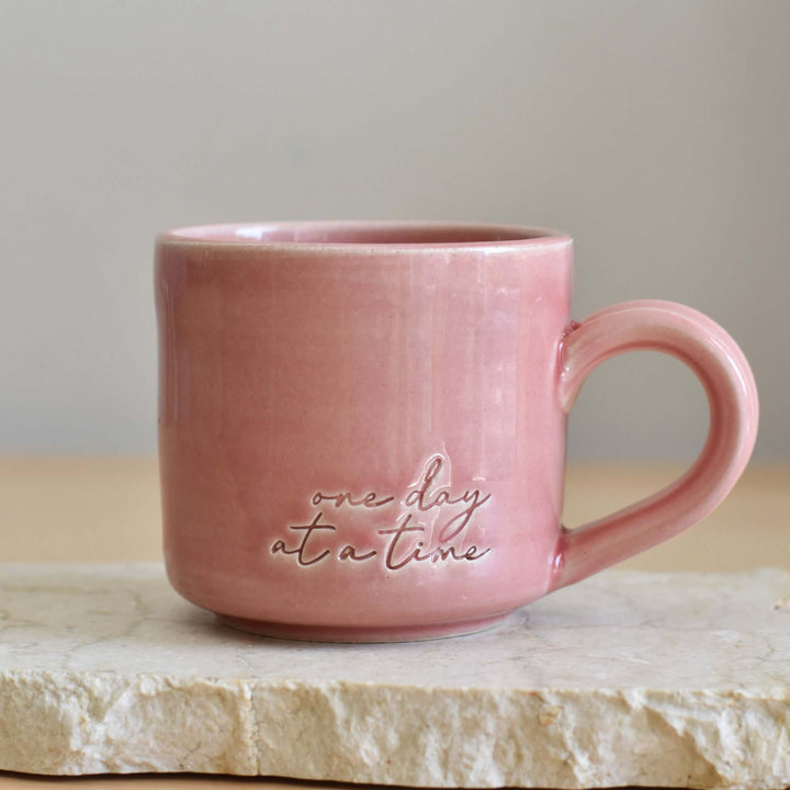 One Day at a Time Mug, pink ceramic mug with hand-glazed finish, features embossed text, sitting on a stone surface.