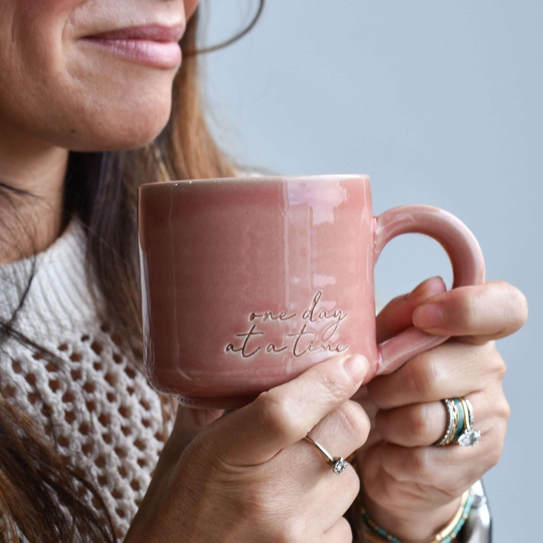 One Day at a Time Mug in pink pottery, featuring embossed text, held by a person in a knit sweater, with rings on fingers.