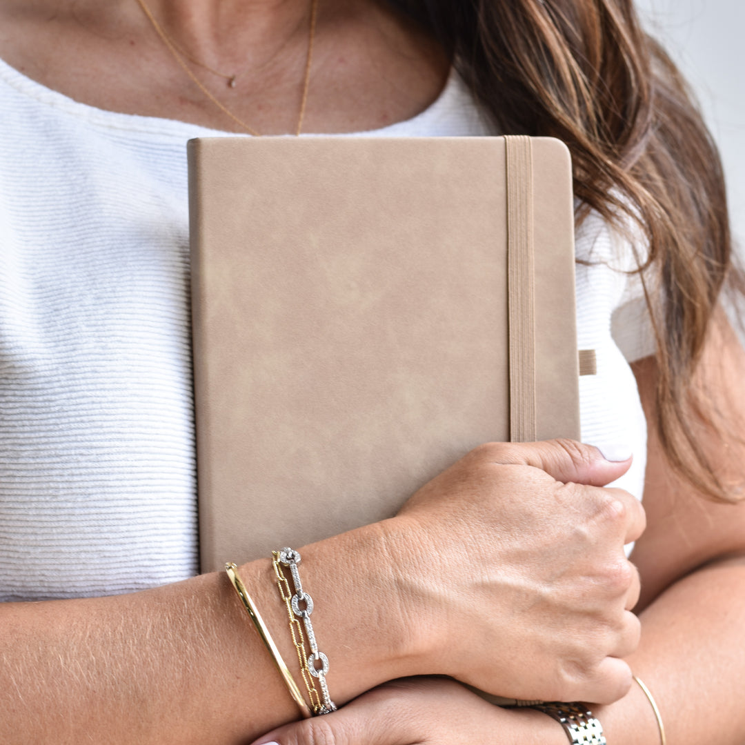 A close-up lifestyle shot of a person holding a tan hardcover notebook, wearing gold and silver chain bracelets on one wrist and a white ribbed top.