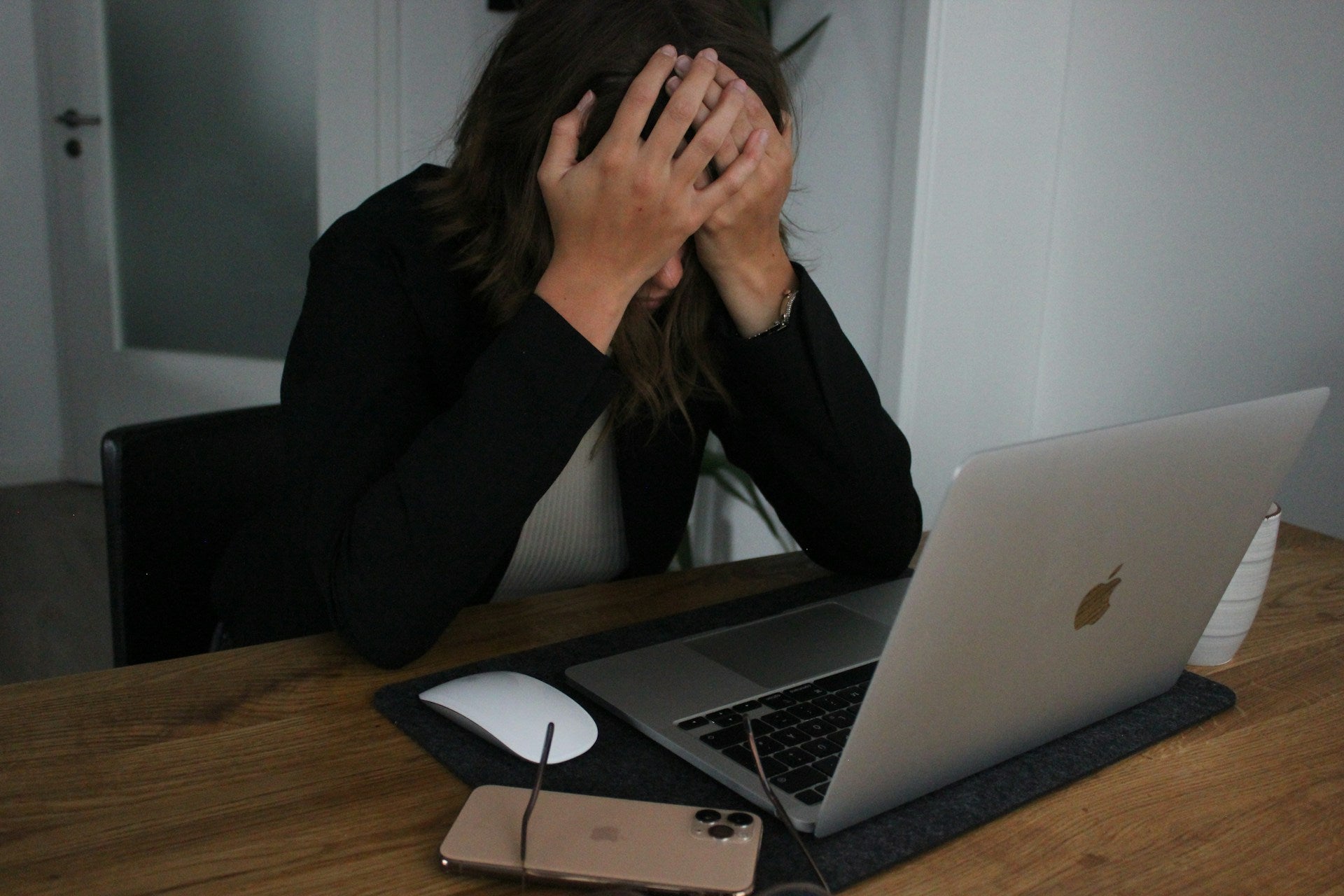photo of a woman sat at a desk with her hands covering her eyes, sitting in front of a laptop with a white computer mouse, iphone and pair of glasses sitting next to the laptop