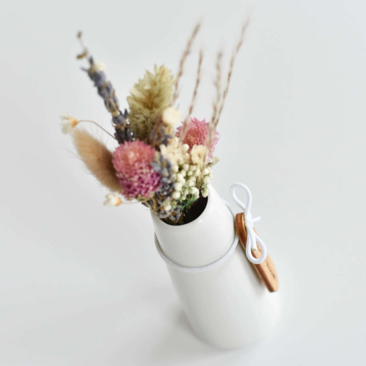 Memories Bloom Forever Dried Floral Bouquet in a white bud vase with dried pink, purple, and beige flowers and an engraved wooden tag.