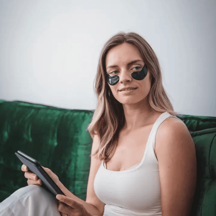 Young woman with black hydrogel eye masks relaxes on a green velvet couch, holding a tablet. Soothing Under Eye Hydrogel Mask.