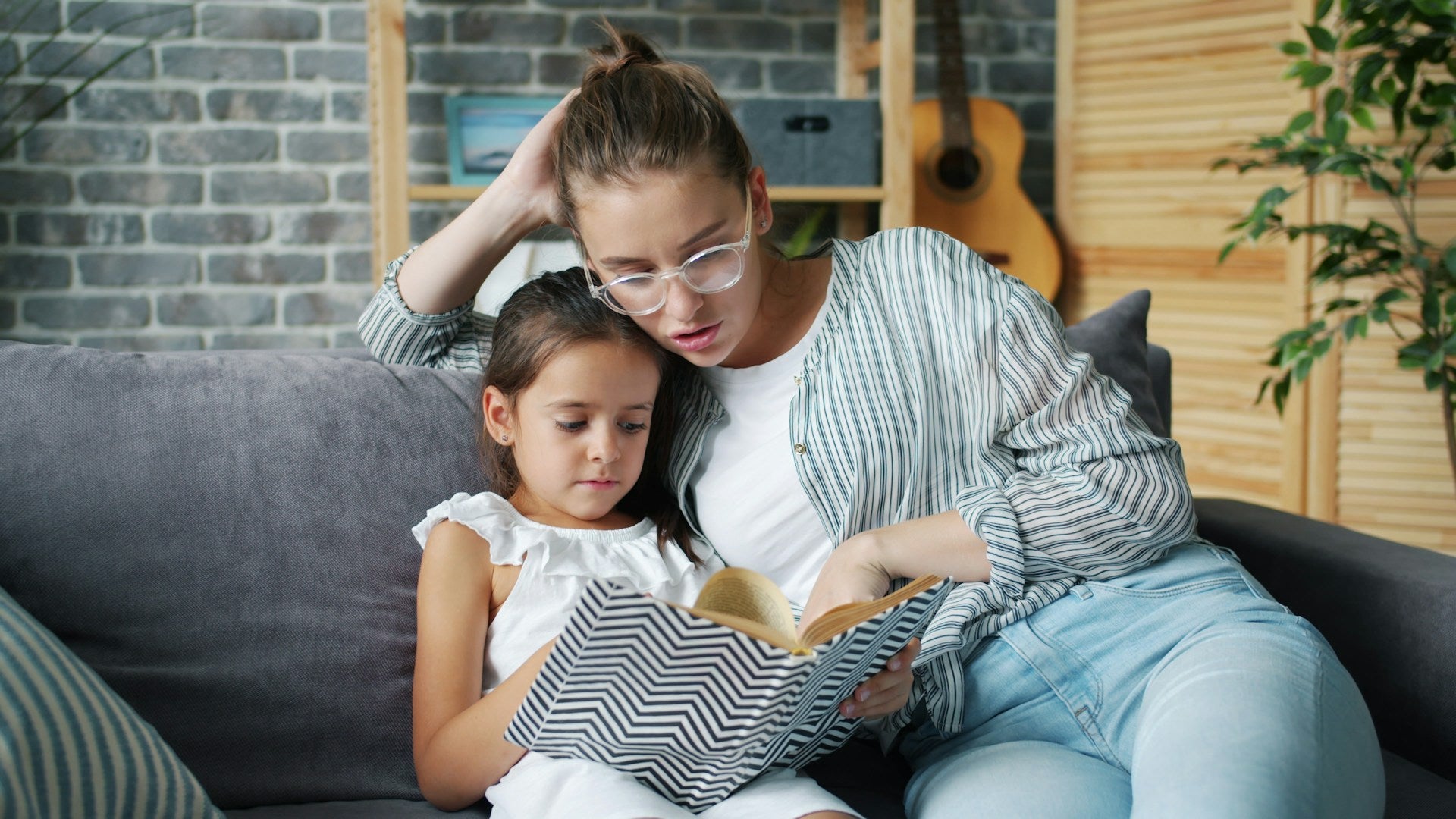 a woman in striped button down shirt sitting with a young girl in a white dress on a couch, both looking a the pages of a book
