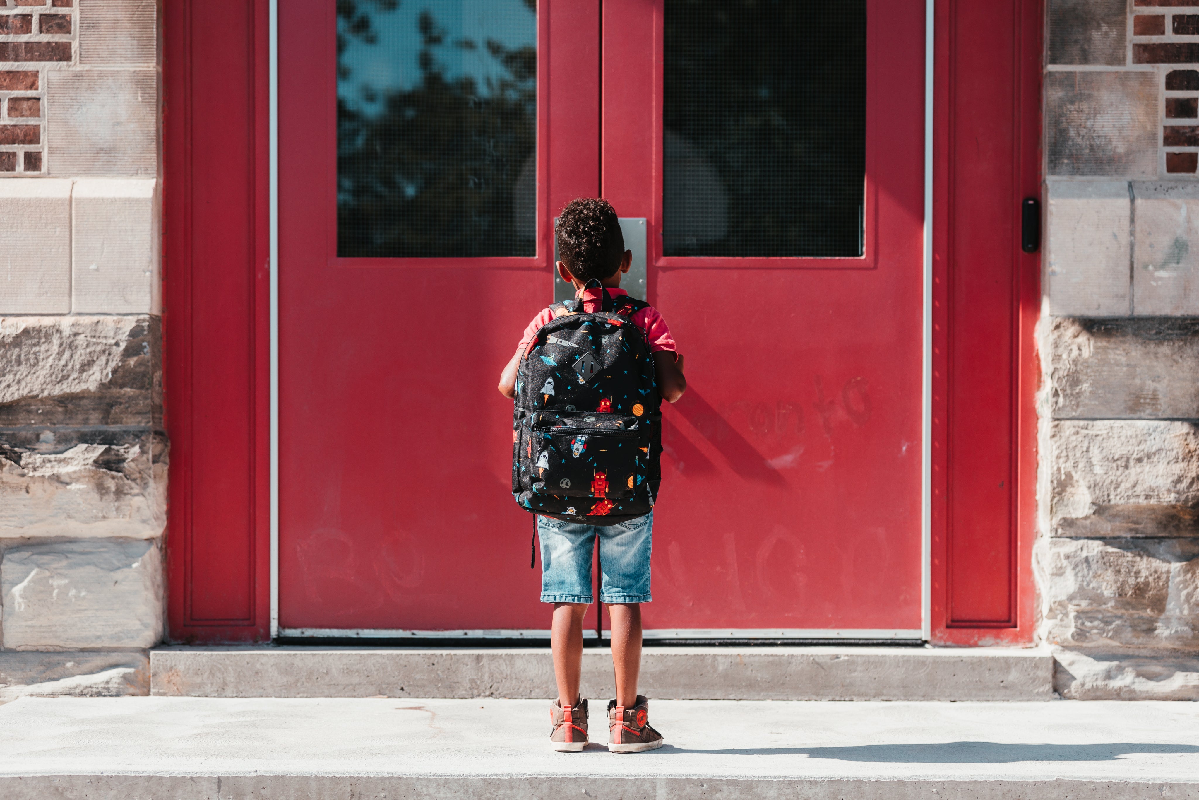 photo of a child wearing a black backpack with robots and rockets on it, standing in front of two red doors