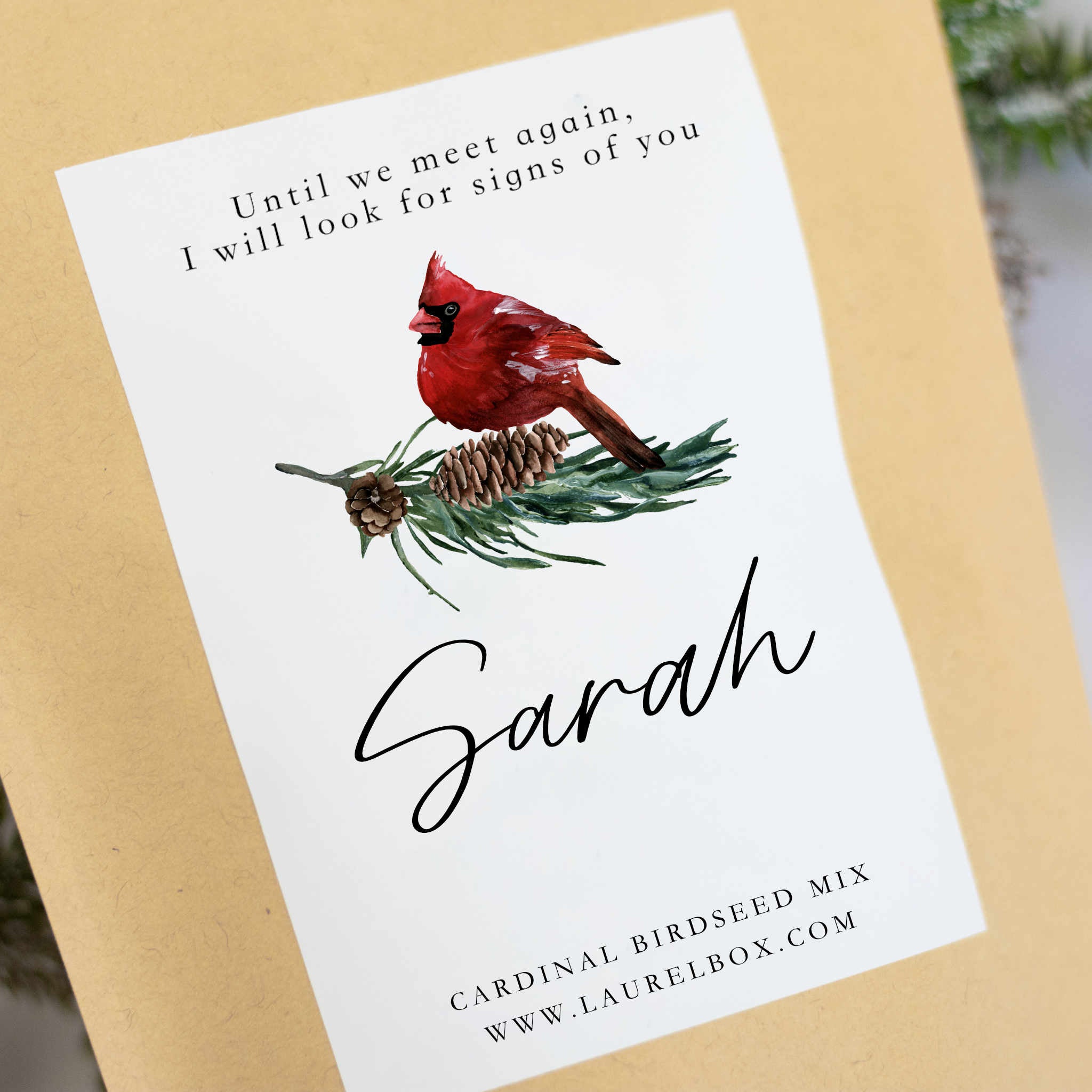 A close-up of a personalized gold cardinal seed packet is angled on top of pine branches in the background. The packet features the text "Until we meet again, I will look for signs of you", a cardinal symbol, the text "Sarah," and "Cardinal Birdseed Mix" at the bottom.