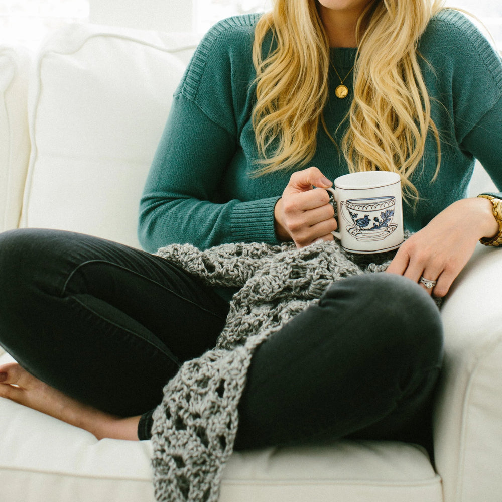 A close-up of a woman with blonde hair wearing a green sweater and jeans is sitting on a white couch, holding a coffee mug, and having a prayer shawl on her lap.