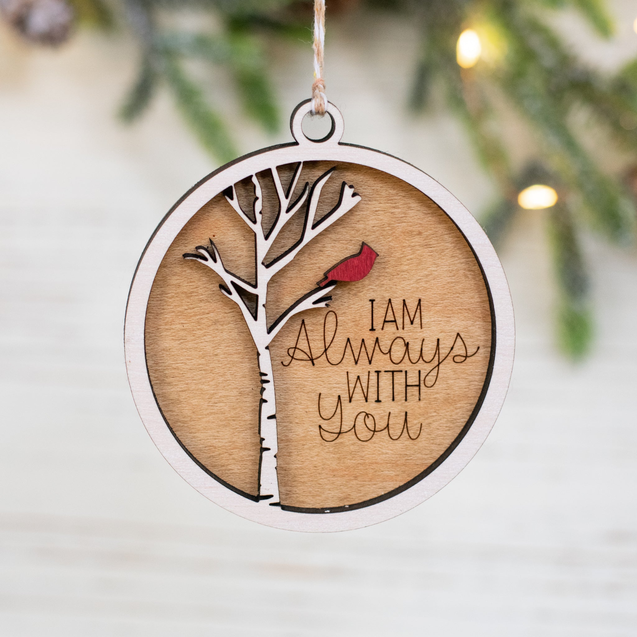 A close-up of a maple wood ornament featuring a cardinal on a white tree, with the text "I am always with you" engraved and hanging on a blurred Christmas tree in the background. 