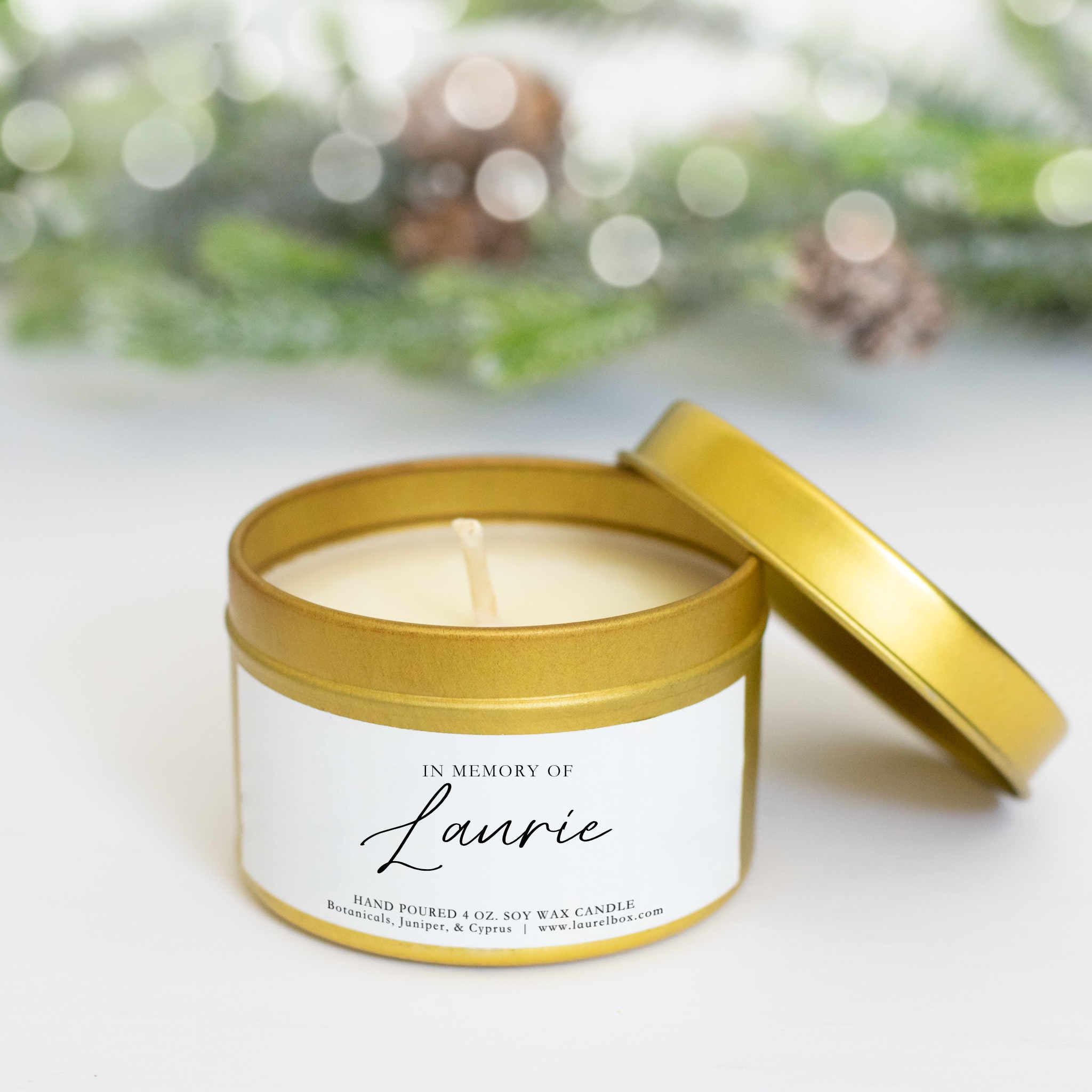 A close-up of a gold candle with a In Memory of Laurie sticker in the middle with the lid resting on the right