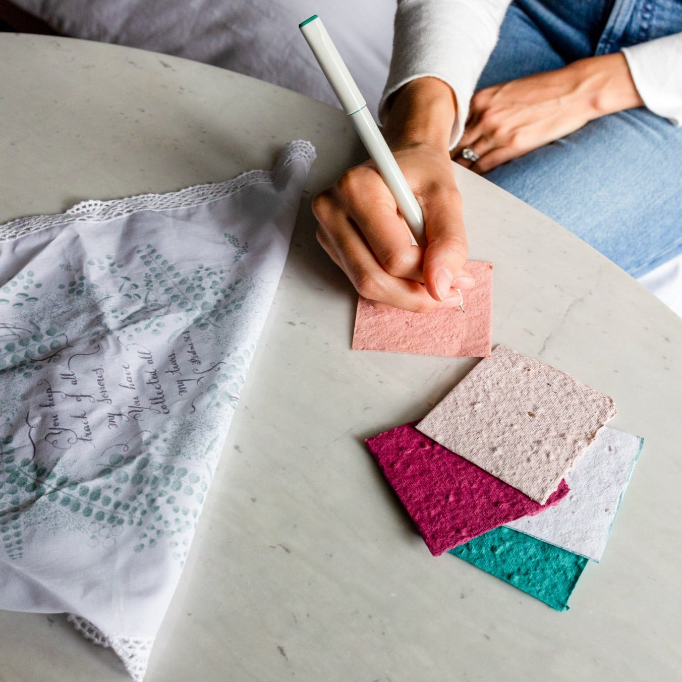 photo of a hand holding a pen writing on a piece of plantable seed paper with other seed paper pieces next to it and a tea towel laying nearby