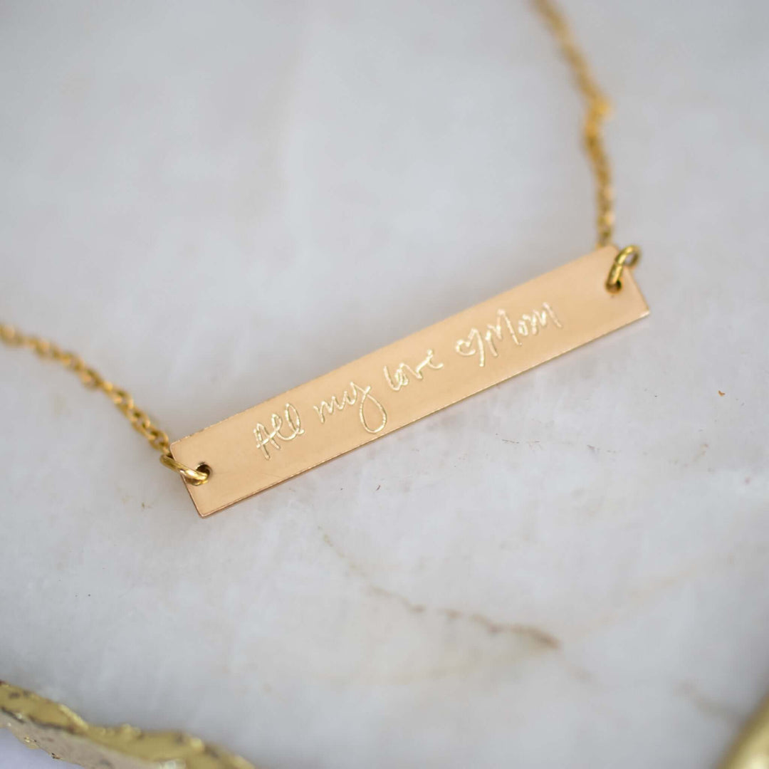 Custom Engraved Handwriting Necklace on a white marble surface. Gold bar charm with engraved text, attached to a gold chain.