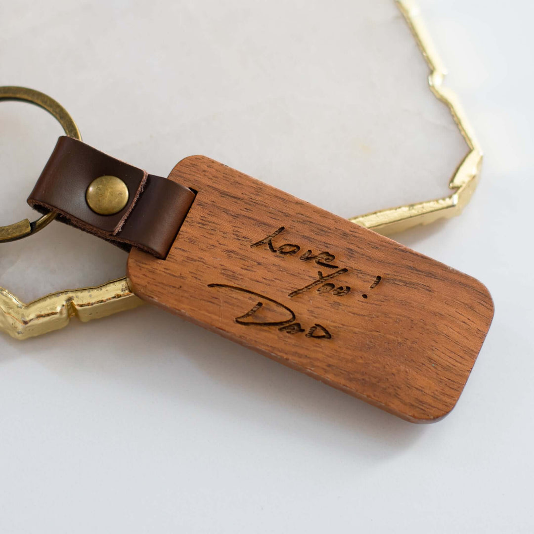 Custom Keychain with Wooden Charm featuring engraved text on brown wood, attached with a brown leather strap and gold keyring.