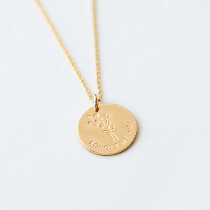 Gold necklace featuring a round pendant with an engraved forget-me-not flower and the text "FORGET ME NOT," on an 18" chain.
