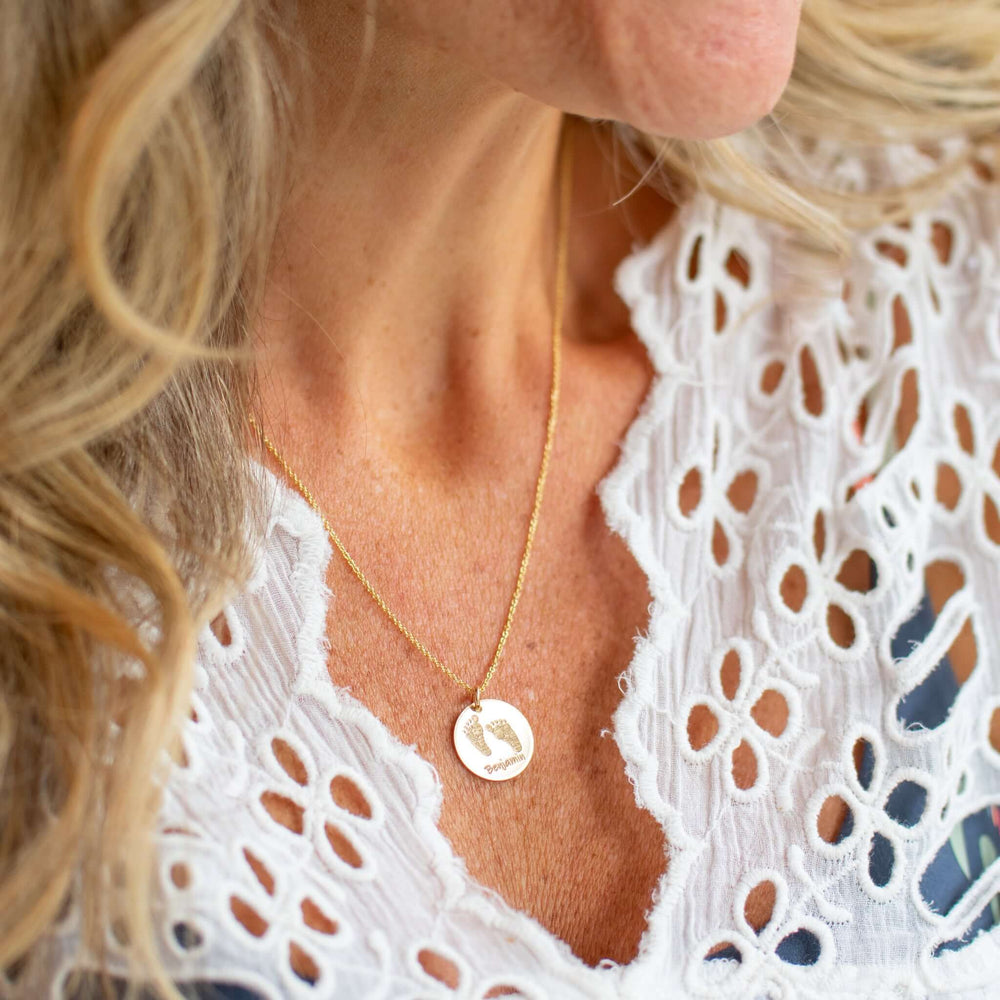 Custom Handprint or Footprint Necklace with engraved footprints on a gold pendant, worn on a woman with a white embroidered top.