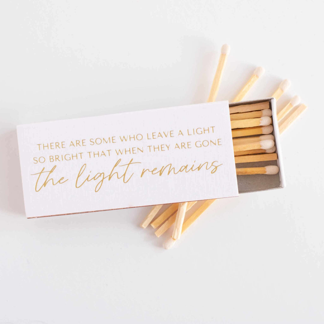 Their Light Remains Matches in a white linen box with gold foil text; inside are wooden matches with white tips.