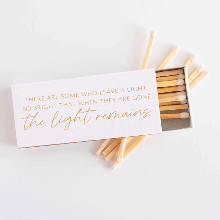 Their Light Remains Matches in a white linen box with gold foil text; inside are wooden matches with white tips.