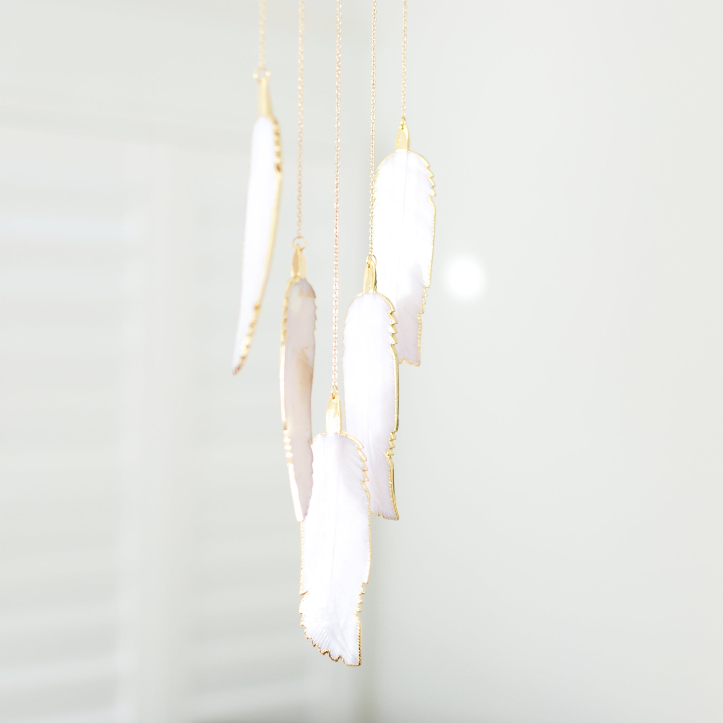 A close-up of a wind chime hanging against a white wall features five mother-of-pearl-shaped feathers
